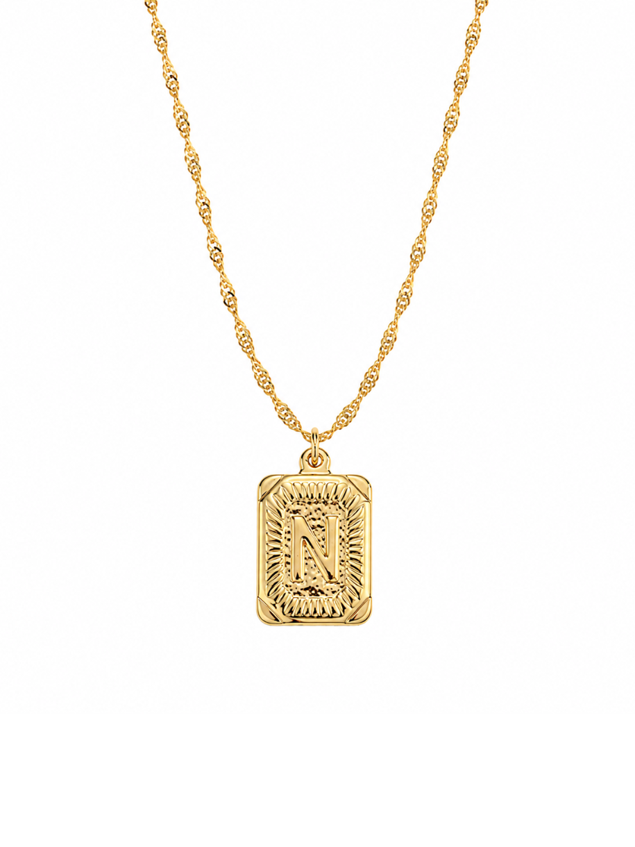 The Vintage Initial Necklace features a rectangular gold pendant engraved with your chosen initial in a decorative script, framed by an ornate sunburst border. It hangs from a twisted gold rope chain.