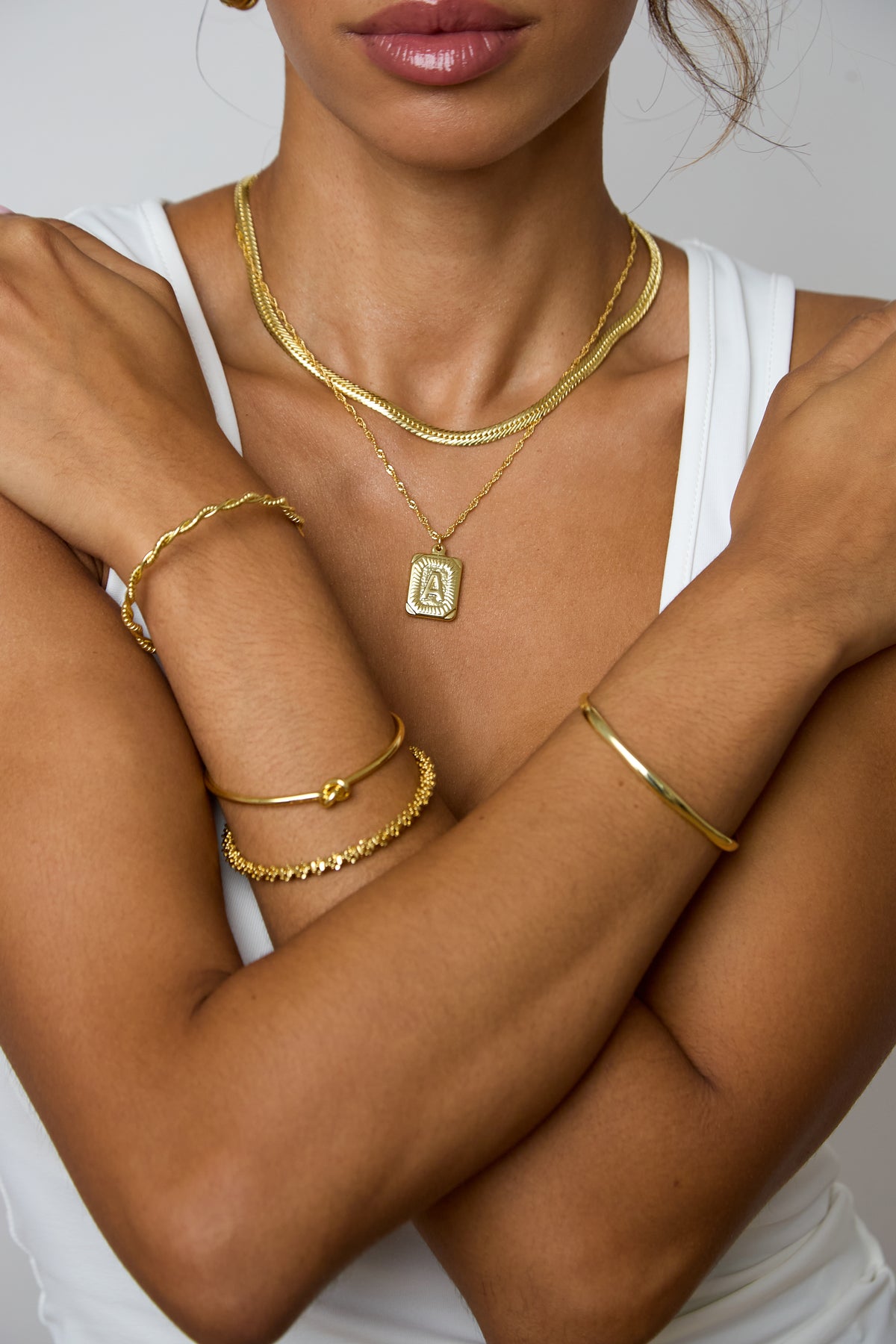 A close-up of a model wearing multiple gold jewelry pieces, including a layered necklace with a square pendant and several bracelets on both wrists, styled with a white tank top against a neutral gray background.
