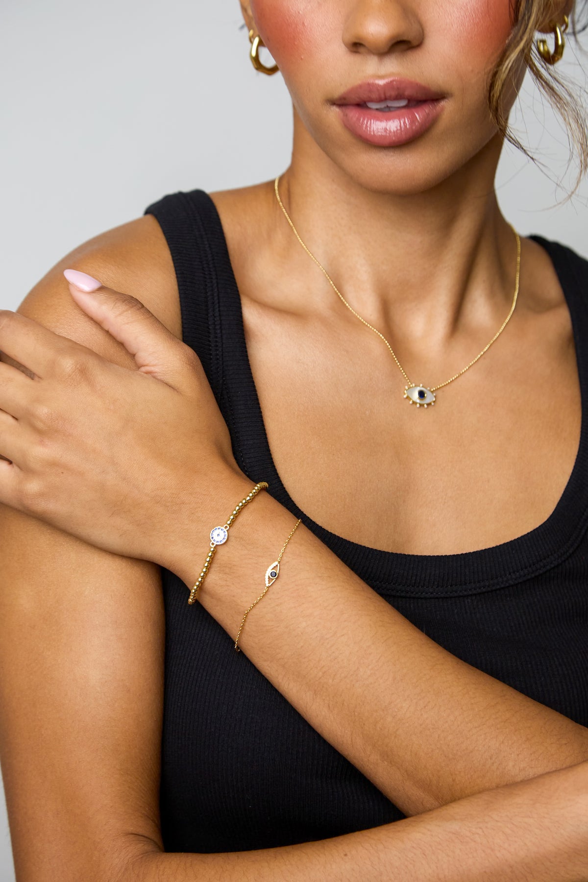 A close-up lifestyle shot of a model wearing a delicate gold jewelry set, including an eye-shaped pendant necklace, a matching bracelet, and hoop earrings, styled with a black ribbed tank top against a neutral gray background.