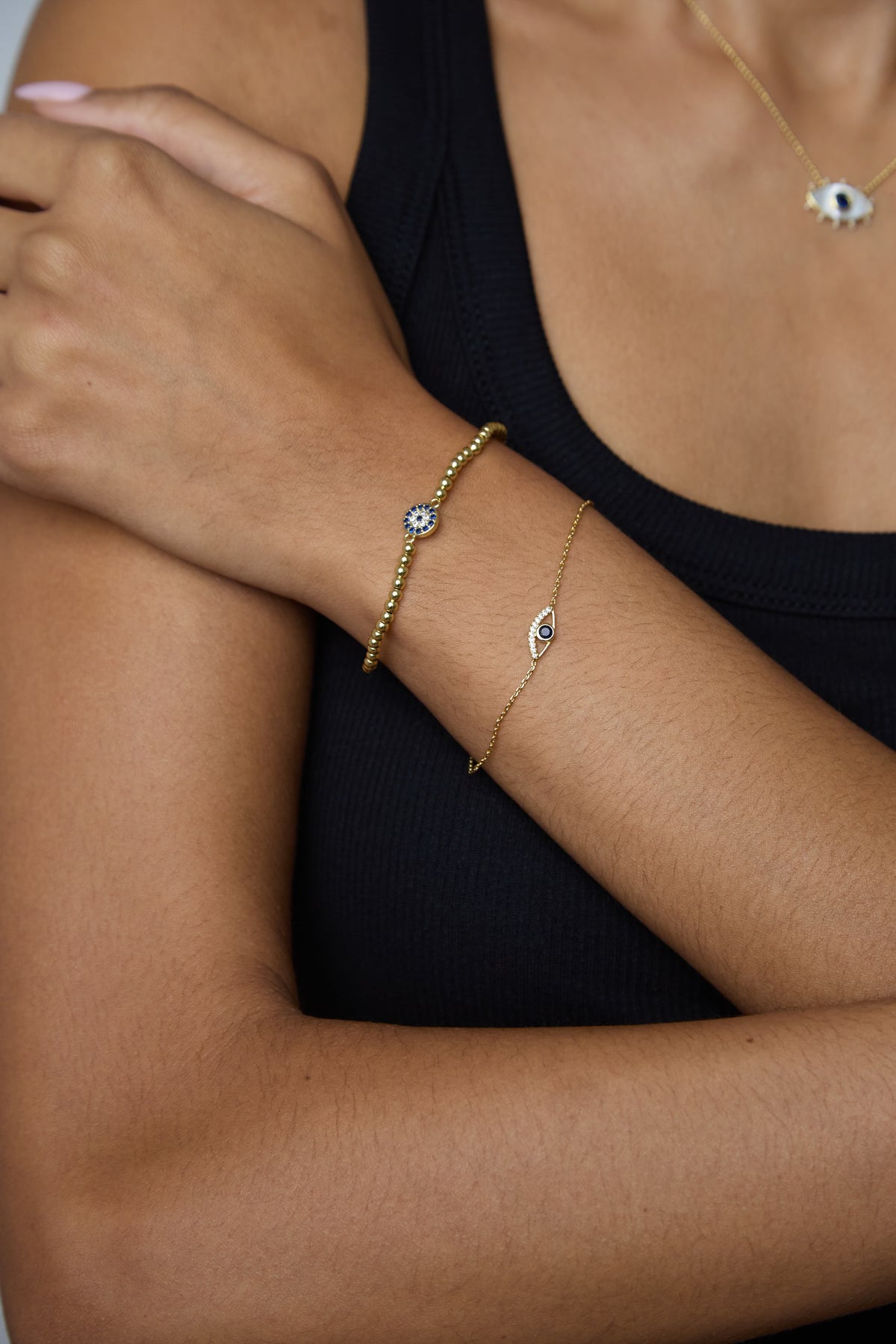 A close-up of a person's arm wearing two delicate gold bracelets, one with a small evil eye charm and the other with a leaf charm, both accented with blue stones, worn over a black ribbed tank top.