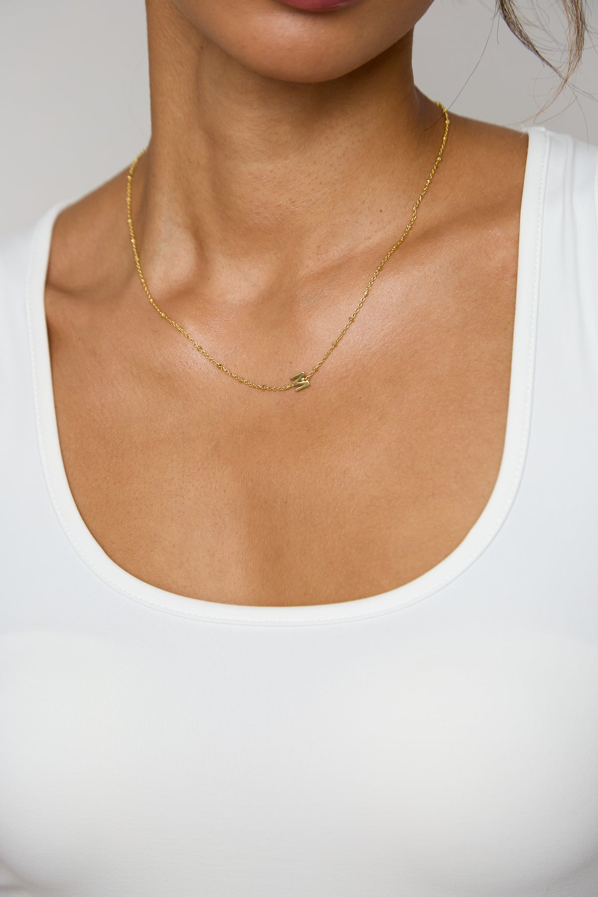 A close-up of a model wearing a delicate gold chain necklace with a small, subtle pendant, styled with a white scoop-neck top against a neutral gray background.
